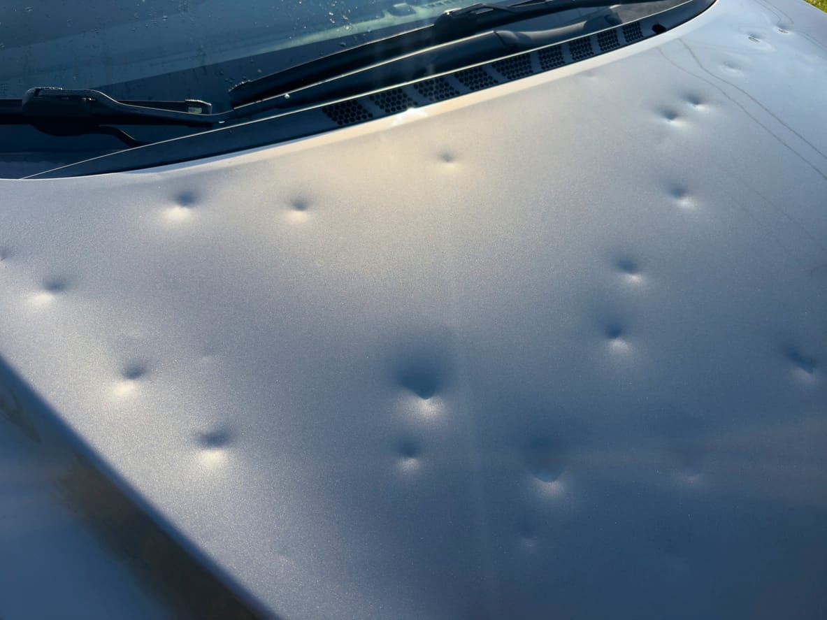Silver car hood covered in shallow round dents from hail